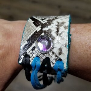 May include: A black and white snakeskin bracelet with a turquoise and black tassel. The bracelet has a large, clear rhinestone in the center.