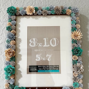 May include: A brown picture frame with a white mat and a wood nameplate that reads "Lydia Wren". The frame is decorated with paper flowers in shades of teal, beige, and gray. The frame is labeled with the sizes "8x10" and "5x7" in inches and centimeters.