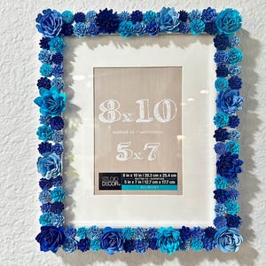 May include: White picture frame with a blue paper flower border. The frame is for 8x10 inch photos and has a 5x7 inch mat. The frame is labeled "Studio Decor" and "Belmont".