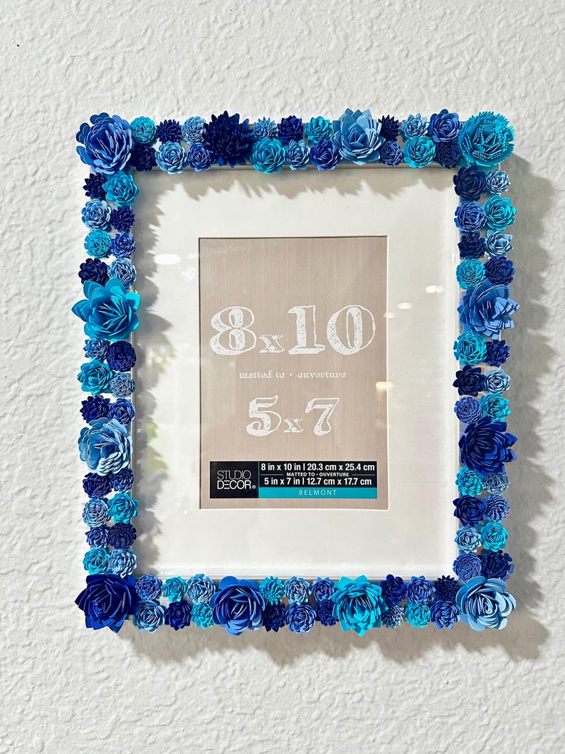 May include: White picture frame with a blue paper flower border. The frame is for 8x10 inch photos and has a 5x7 inch mat. The frame is labeled "Studio Decor" and "Belmont".