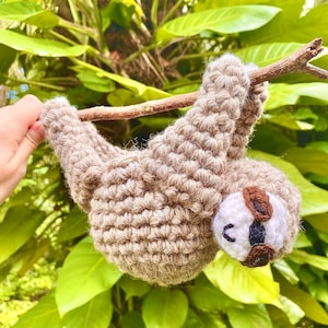 May include: A crocheted sloth toy hanging from a branch. The sloth is brown with a white face and black eyes. It is wearing brown glasses.