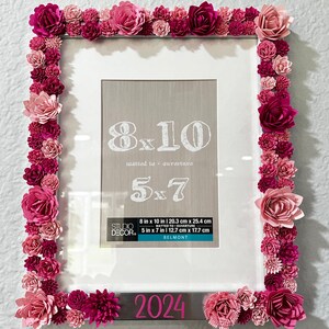 May include: A white picture frame with a silver border decorated with pink and red paper flowers. The frame is an 8x10 size, matted to a 5x7 size. The frame has the text "2024" on the bottom.
