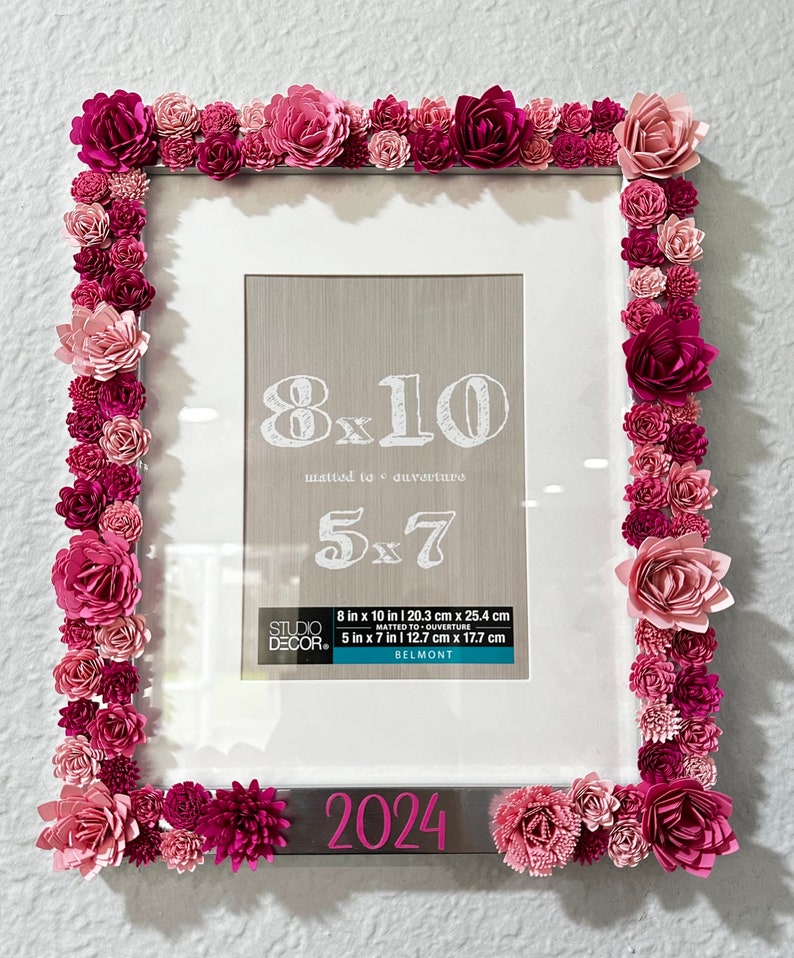 May include: A white picture frame with a silver border decorated with pink and red paper flowers. The frame is an 8x10 size, matted to a 5x7 size. The frame has the text "2024" on the bottom.
