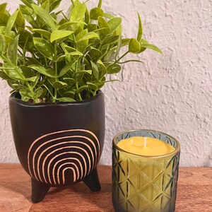 May include: A yellow candle with a single wick in a green glass jar with a geometric pattern. The jar is sitting on a wooden surface next to a black planter with a white rainbow design.