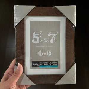 May include: A brown wood picture frame with a white mat and a silver border. The frame is 5x7 inches and can be used to display a 4x6 inch photo. The frame is labeled "Studio Decor" and "Belmont".