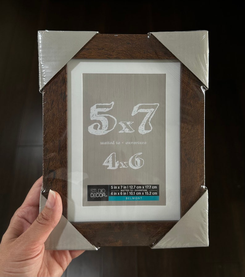 May include: A brown wood picture frame with a white mat and a silver border. The frame is 5x7 inches and can be used to display a 4x6 inch photo. The frame is labeled "Studio Decor" and "Belmont".