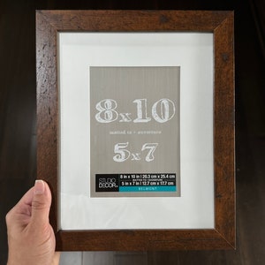 May include: A brown wood picture frame with a white mat. The frame is 8 inches by 10 inches and can be used to mat a 5 inch by 7 inch photo. The frame is labeled "Studio Decor" and "Belmont".