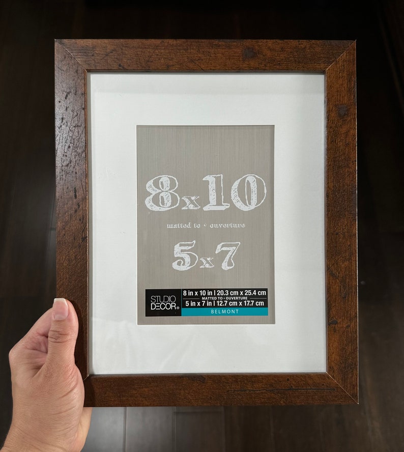 May include: A brown wood picture frame with a white mat. The frame is 8 inches by 10 inches and can be used to mat a 5 inch by 7 inch photo. The frame is labeled "Studio Decor" and "Belmont".