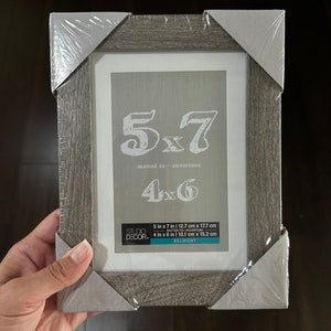 May include: A gray wood picture frame with a white mat. The frame is labeled "5x7 matted to ouverture 4x6". The frame is 5 inches by 7 inches and can fit a 4 inch by 6 inch photo.