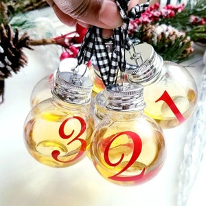 May include: Clear glass ornaments filled with a yellow liquid, each with a red number. The ornaments have silver caps and are tied together with a black and white checkered ribbon. Festive holiday decor.