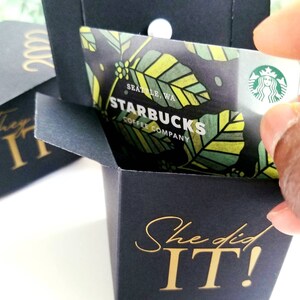 K&ouml;nnte beinhalten: Eine schwarze Geschenkbox mit einer goldenen Aufschrift, die "She did IT!" lautet. Die Box ist ge&ouml;ffnet und eine Starbucks-Geschenkkarte befindet sich im Inneren. Die Geschenkkarte hat ein gr&uuml;nes, gelbes und braunes Design mit dem Starbucks-Logo und dem Text "Starbucks Coffee Company" in Gold.