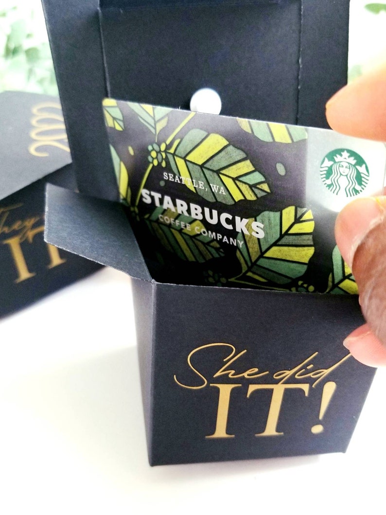 K&ouml;nnte beinhalten: Eine schwarze Geschenkbox mit einer goldenen Aufschrift, die "She did IT!" lautet. Die Box ist ge&ouml;ffnet und eine Starbucks-Geschenkkarte befindet sich im Inneren. Die Geschenkkarte hat ein gr&uuml;nes, gelbes und braunes Design mit dem Starbucks-Logo und dem Text "Starbucks Coffee Company" in Gold.