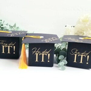 K&ouml;nnte beinhalten: Drei schwarze Boxen in Form von Abschlusskappen mit goldenen Schriftz&uuml;gen. Die Boxen sagen "They did it!" "He did it!" und "She did it!" Die Boxen haben gelbe Quasten.