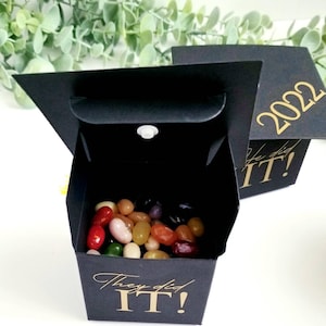 K&ouml;nnte beinhalten: Schwarze Box in Form einer Abschluss-Kappe mit goldener Aufschrift "They did IT!" und dem Jahr "2022". Die Box ist mit bunten Gummib&auml;rchen gef&uuml;llt.
