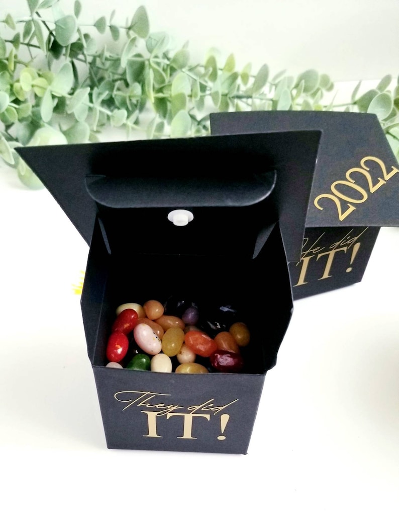 K&ouml;nnte beinhalten: Schwarze Box in Form einer Abschluss-Kappe mit goldener Aufschrift "They did IT!" und dem Jahr "2022". Die Box ist mit bunten Gummib&auml;rchen gef&uuml;llt.