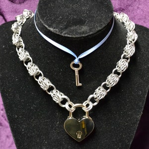 May include: A silver chain necklace with a heart-shaped padlock and a small key charm. The necklace is on a black velvet display stand. The chain is made of interlocking metal rings.
