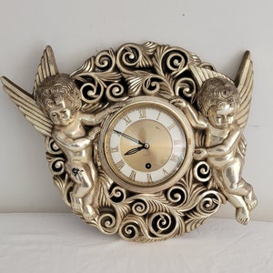 May include: A gold-toned wall clock with a circular face and Roman numeral markings. Two cherubs with wings are flanking the clock, which is set against a background of swirling, ornate designs.
