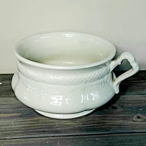 May include: White ceramic chamber pot with a decorative design around the rim and two handles. The pot is sitting on a dark wooden surface.