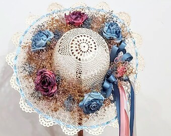 May include: A white crocheted hat with blue and pink ribbons and floral embellishments. The hat is displayed on a wooden stand with a heart-shaped base.