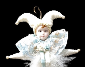 May include: A porcelain doll dressed as a jester with a white and blue costume, white feathers, and a blue ribbon. The doll has a white face with painted features and brown hair.