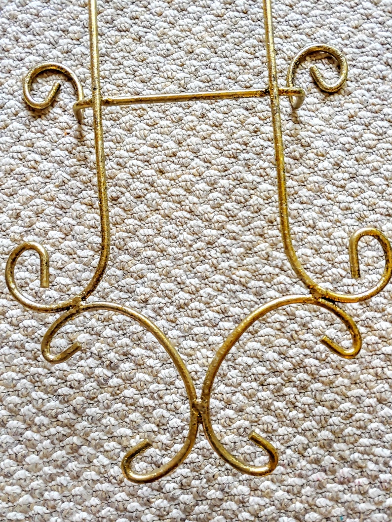 May include: A gold metal plate stand with a decorative design. The stand has a rectangular shape with a curved bottom and two decorative loops at the top.