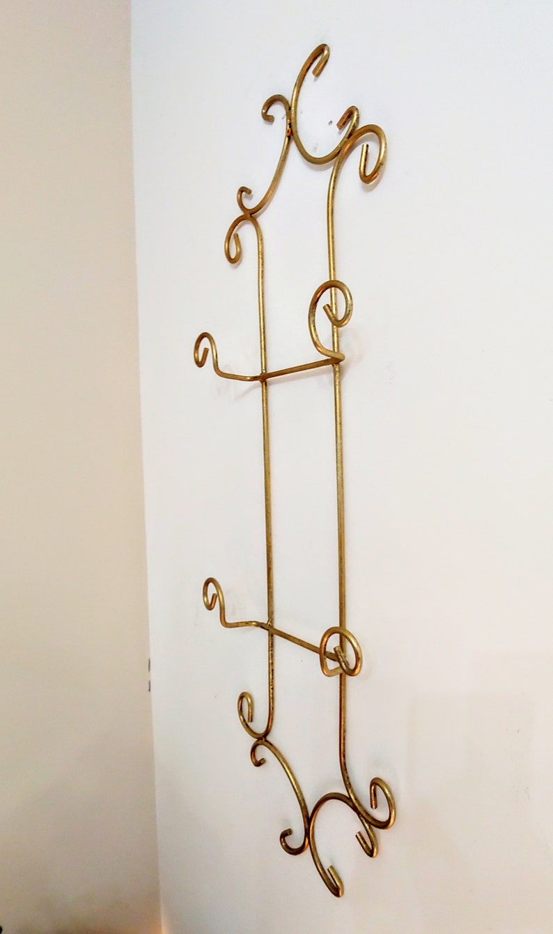 May include: Gold metal wall mounted plate rack with decorative scrollwork. The rack has three tiers for displaying plates.