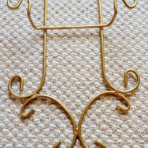 May include: Gold metal plate stand with decorative scrollwork. The stand has two hooks at the top and a decorative base.