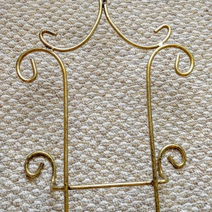 May include: A gold metal stand with a decorative design. The stand has a rectangular shape with a horizontal bar across the middle. The stand is made of thin metal and has a delicate, ornate design.