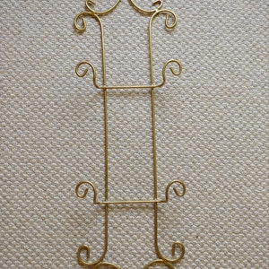 May include: Gold metal plate stand with ornate scrollwork details. The stand has two shelves for displaying plates.