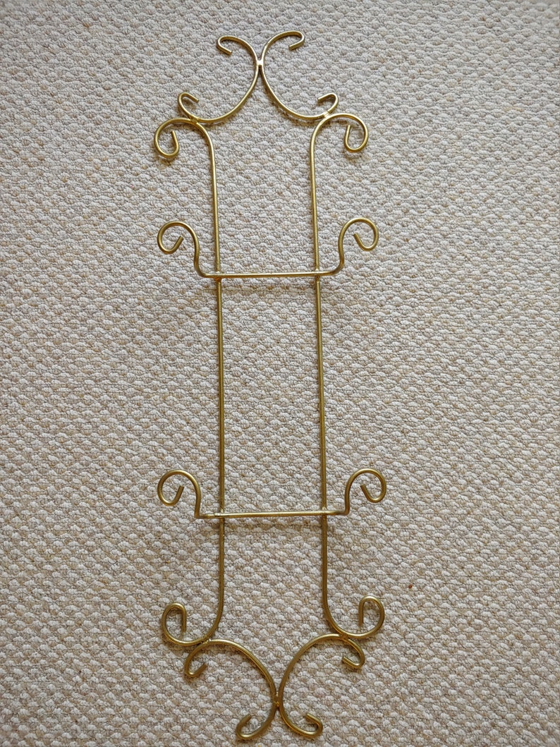 May include: Gold metal plate stand with ornate scrollwork details. The stand has two shelves for displaying plates.