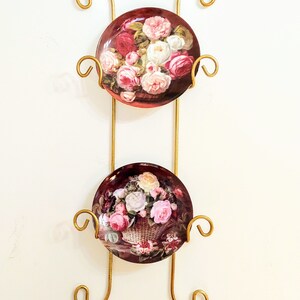 May include: A gold metal plate rack with two decorative plates featuring floral designs. The plates have a dark background with pink and white roses.