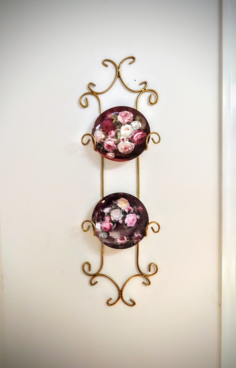 May include: A gold metal plate rack with two decorative plates featuring floral designs. The plates are a dark burgundy color with pink and white roses.