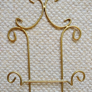 May include: Gold metal stand with a decorative design. The stand has two vertical supports and a horizontal bar with a decorative curl on each end.