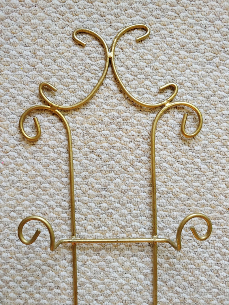 May include: Gold metal stand with a decorative design. The stand has two vertical supports and a horizontal bar with a decorative curl on each end.