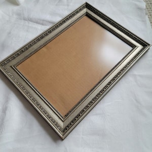 May include: A silver picture frame with an ornate design. The frame is empty and has a brown mat.