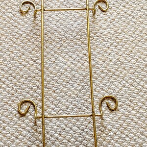 May include: A gold metal decorative stand with four hooks on each side. The stand has a decorative design at the top and bottom.