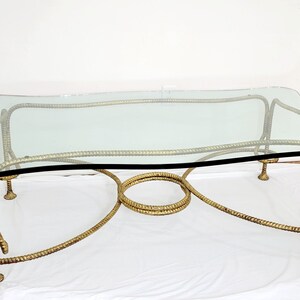 May include: A gold-toned metal coffee table with a glass top. The base of the table is made of twisted metal that resembles rope.
