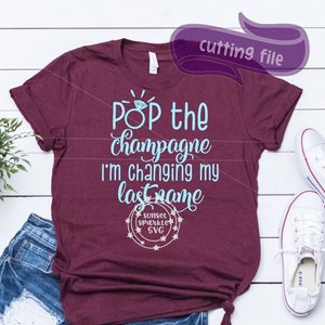 Könnte beinhalten: Burgunderfarbenes T-Shirt mit dem Text "Pop the champagne I'm changing my last name" in Hellblau. Das Shirt wird mit Jeansshorts und weißen Turnschuhen kombiniert. Links ist eine kleine Pflanze.