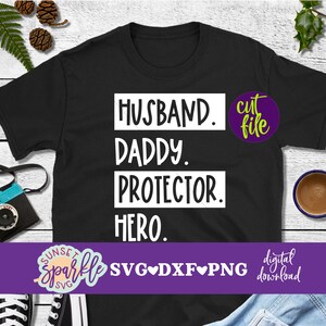 May include: Black t-shirt with white text that reads "Husband. Daddy. Protector. Hero." The text is in a box with rounded corners. The shirt is on a wooden surface with a camera, a cup of coffee, and a pair of trainers.