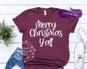May include: A maroon t-shirt with the text "Merry Christmas y'all" in white lettering. The shirt is laying on a white wooden surface with a pair of white sneakers and denim shorts.
