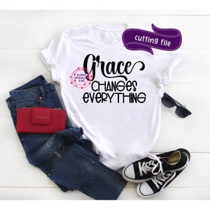 May include: White t-shirt with the text "Grace Changes Everything" in black. The shirt is laid out on a white surface with blue jeans, a red wallet, sunglasses, and black trainers.