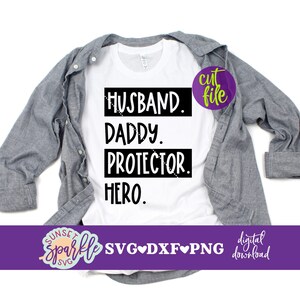 May include: A white t-shirt with black text that reads "Husband. Daddy. Protector. Hero." The shirt is folded and laid on a grey surface.