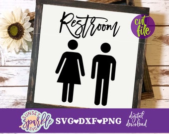 May include: A white sign with black text that reads "Restroom" with a male and female symbol underneath. The sign is framed in a dark wood frame and is sitting on a white wood surface.