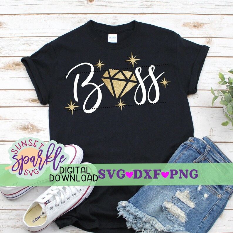 Puede incluir: Una camiseta negra con la palabra "Boss" escrita en cursiva blanca con un diamante dorado en el medio de la palabra. El diamante tiene un efecto brillante. Hay estrellas doradas alrededor de la palabra. La camiseta est&aacute; sobre una superficie de madera con un par de zapatillas blancas y un par de pantalones cortos de mezclilla azul.