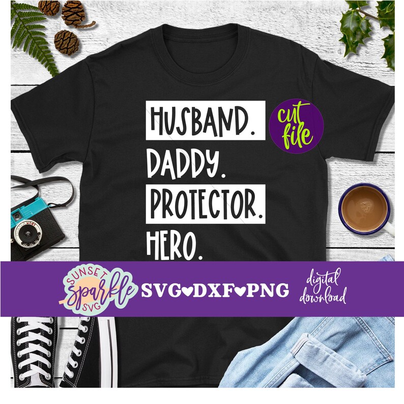 May include: Black t-shirt with white text that reads "Husband. Daddy. Protector. Hero." The text is in a box with rounded corners. The shirt is on a wooden surface with a camera, a cup of coffee, and a pair of trainers.