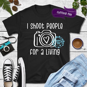 May include: Black t-shirt with white text that reads "I shoot people for a living". A white camera with a heart in the lens is pictured above the text.
