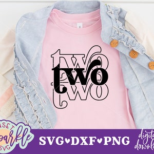 May include: A pink t-shirt with a black graphic that says "two" in a stylized font. The shirt is folded and displayed on a white surface with a denim jacket and a pink bow.