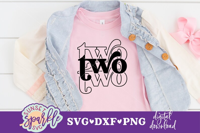 May include: A pink t-shirt with a black graphic that says "two" in a stylized font. The shirt is folded and displayed on a white surface with a denim jacket and a pink bow.