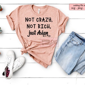 May include: A pink t-shirt with the text "NOT CRAZY, NOT RICH. just Asian" in black lettering. The shirt is laid flat on a white wooden surface with a pair of white sneakers, a pair of blue jeans, and small potted plants.