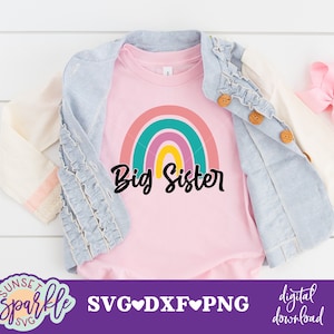 May include: A pink t-shirt featuring a rainbow and the text "Big Sister". The shirt is displayed on a white surface with a denim jacket and a pink bow.